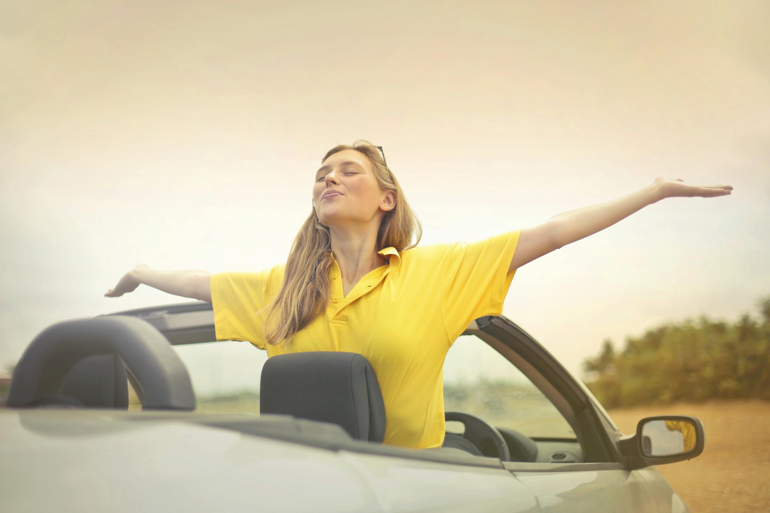 A cheerful woman in a yellow shirt enjoys freedom on a sunny drive in a convertible.